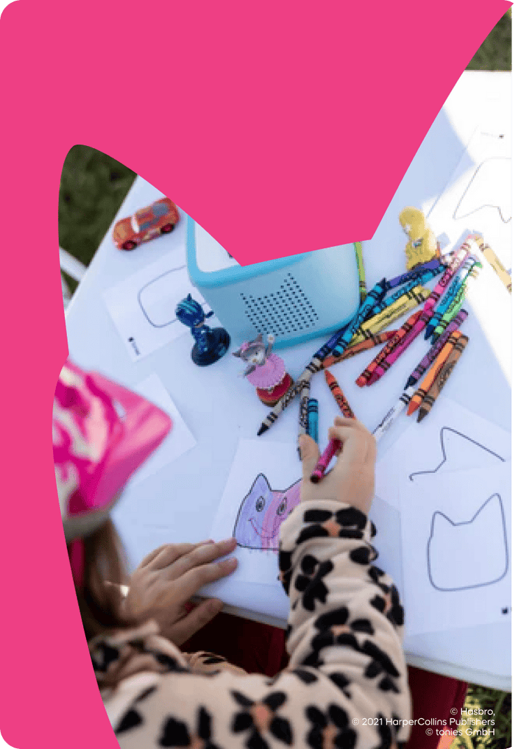 Child coloring with crayons on a table outdoors, listening to a blue Toniebox 2