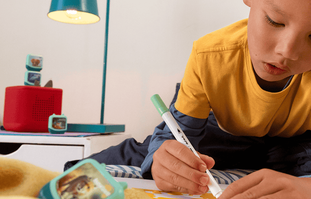 child coloring on a bed while listening to pocket tonies on a red toniebox 2