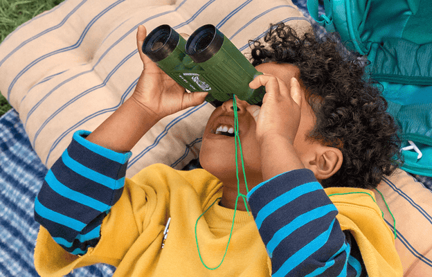 Child using binoculars on a grassy field with a Listen and Go Bag and cushions nearby