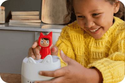 A smiling child in a yellow knit sweater holds a small red hooded figurine above a Toniebox Lite